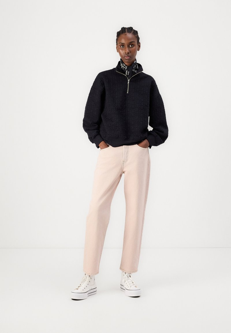 Suéter negro texturizado con cuello alto y cremallera, combinado con pantalones rectos rosa pálido y zapatillas de deporte blancas de caña alta.