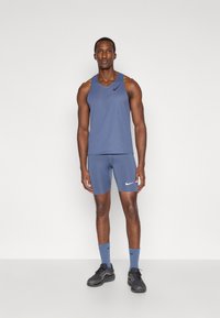 Tenue de sport pour homme comprenant un débardeur sans manches bleu avec texture perforée et un logo blanc, associé à un short bleu assorti et des baskets noires.