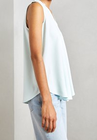 Haut sans manches bleu clair avec col en V, coupe décontractée et forme légèrement évasée, associé à un jean en denim clair. Tissu lisse et design minimaliste.