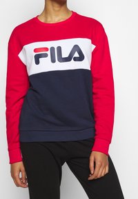 Persoon draagt een rode, witte en marineblauwe FILA-trainingsbroek met een zwarte broek, staat met één hand ontspannen langs het lichaam.