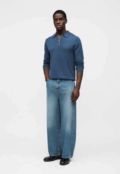 Άνδρας στέκεται φορώντας μπλε πουκάμισο με φερμουάρ και μακριά μανίκια, φαρδιά ξεβαμμένα τζιν και μαύρα παπούτσια, μπροστά σε λευκό φόντο.