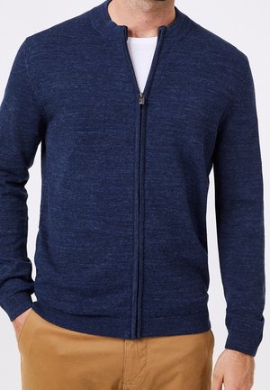 Hombre con un suéter azul marino con cremallera sobre una camisa blanca y pantalones marrón claro, con las manos parcialmente en los bolsillos, contra un fondo liso.