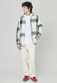 Chemise à boutons avec un motif écossais vert et gris, t-shirt blanc, pantalon crème et baskets rouges. Texture subtile dans le tissu.