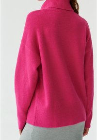 Duży różowy sweter typu pulower z wysokim kołnierzem, ściągaczami przy rękawach i u dołu. Miękka tekstura i luźny fason, pokazany od tyłu.