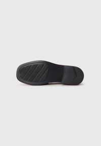 Semelle de chaussure en caoutchouc noir avec motif de grip texturé et design incurvé. Le talon plat présente une finition lisse et un détail de marque discret.