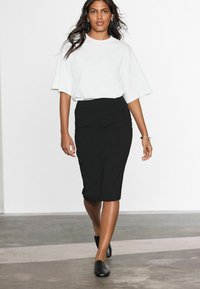 T-shirt blanc surdimensionné et jupe crayon noire ajustée. La jupe a une texture lisse et des détails de ruché subtils. Des chaussures noires à enfiler complètent le look.