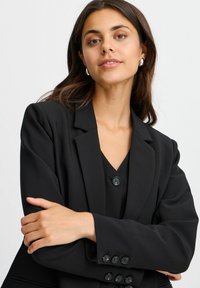 Černý blazer s klopou, zapínáním na jeden knoflík a manžetami na knoflíky. Nosený přes ladící černý top s jemnou texturou.