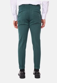 Pantaloni verdi su misura con una texture liscia, dotati di due tasche posteriori con chiusura a bottone e orli risvoltati, abbinati a scarpe nere.