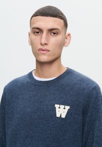 Námornícky modrý pletený sveter s okrúhlym výstrihom a rebrovanými manžetami, na hrudi s bielym logom "M". Hladká textúra.