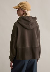 Brauner übergroßer Hoodie mit Kapuze und gerippten Bündchen, mit lockerem Schnitt. Im Hintergrund eine beige Baseballkappe kombiniert.