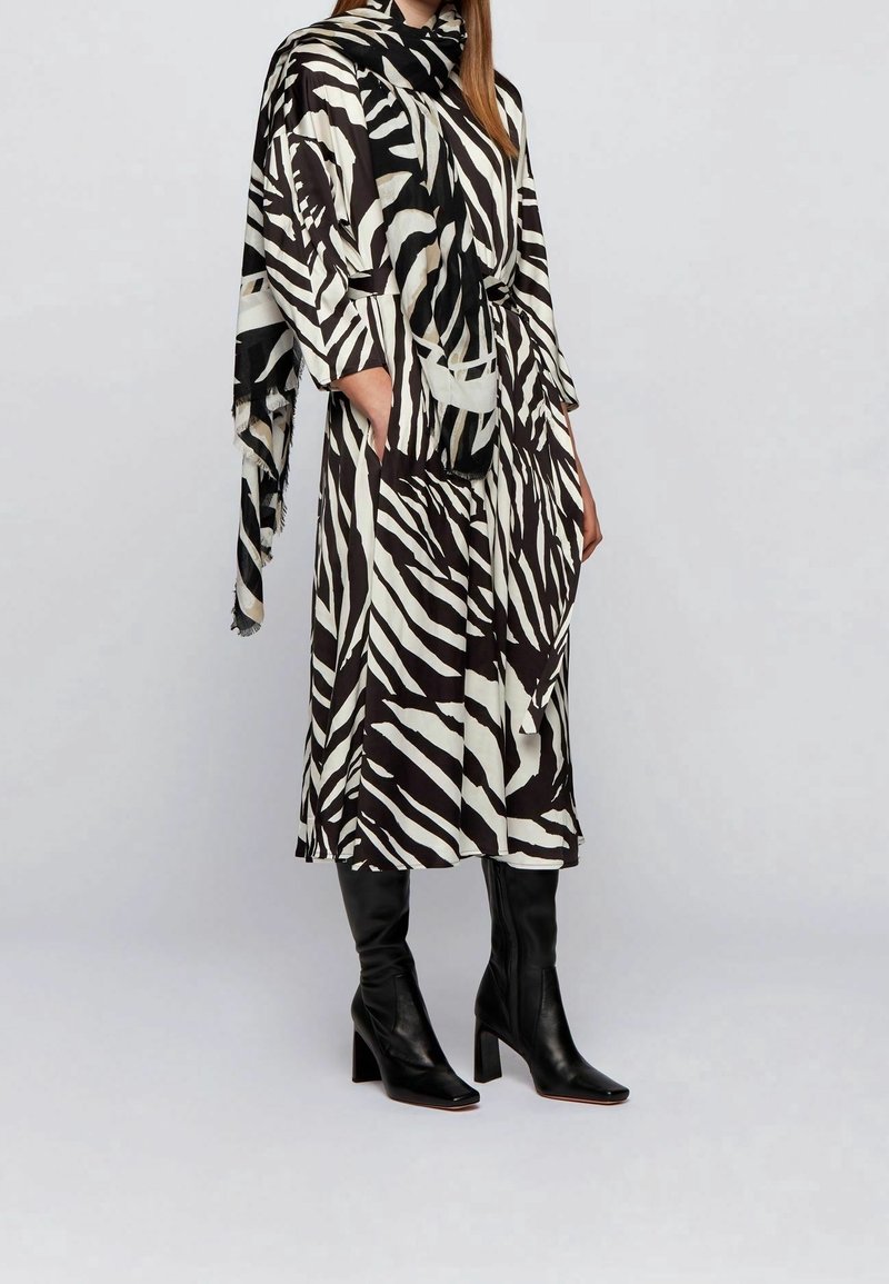 Femme portant une robe midi à rayures zébrées noires et blanches avec une écharpe assortie et des bottes en cuir noires à talons, sur un fond uni.