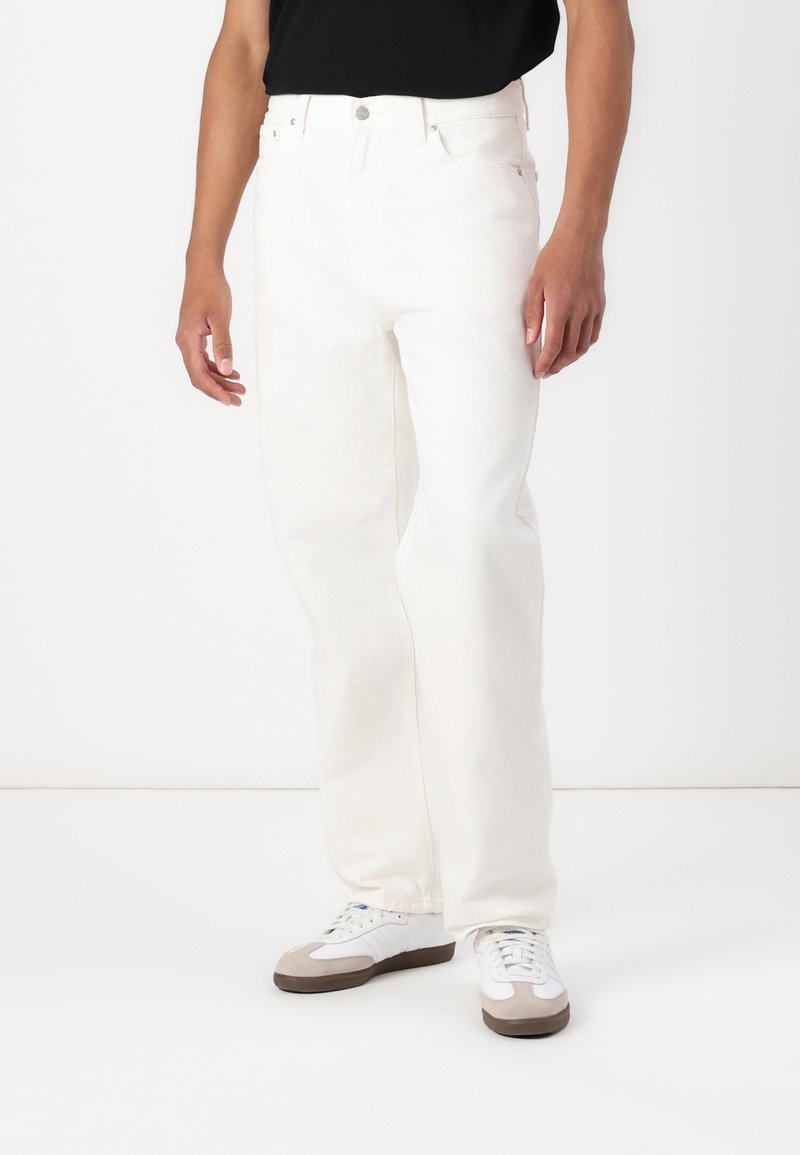 Hvide denim bukser med lige pasform, der har en frontknaplukning og sidelommer, parret med hvide sko og en sort top.