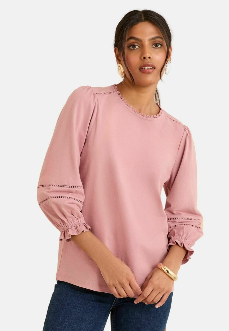 Helle rosa Bluse mit puffigen Ärmeln, Rüschendetails und gestickten Akzenten entlang der Kanten. Kombiniert mit dunkelblauen Jeans und einem goldenen Armband.