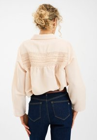 Heledad virsikavärvi pluus, millel on volditud seljaosa ja smock-detailid. Pikad varrukad ja krae; kantud tumehallide teksadega.