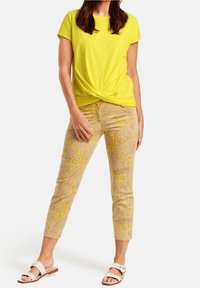 Gul kortärmad topp med knutdetalj fram, ihopparad med beigefärgade byxor med gula blommönster. Vita skosandaler.