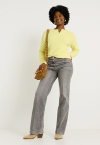 Un pull jaune duveteux avec un design henley, un jean droit gris, un sac bandoulière beige avec des clous et des bottines à talons beige.