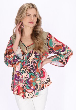 Blondynka w kolorowej, kwiecistej bluzce z oliwkowym koronkowym kołnierzem i białych spodniach, dotykająca kolczyka, spoglądająca na bok, delikatnie się uśmiechająca.