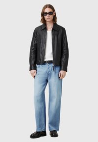 Črna usnjena jakna z zadrgo, bela rebrasta majica, svetlo modri ravni jeans in črne sijoče čevlje. Model nosi temne sončne očala.