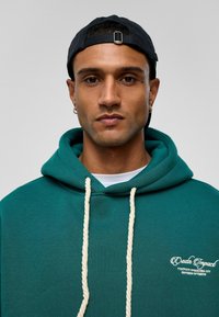Teal hoodie met een voorzak, witte trekkoorden en geborduurde tekst. Draag met een zwarte pet; achtergrond is effen grijs.