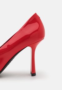 Chaussure à talon haut stilettos rouge brillant avec bout pointu et talon fin et effilé sur un fond blanc uni.