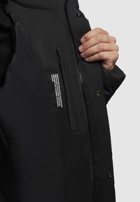 Eine Hand hält eine offene schwarze Jacke und zeigt die Innentasche mit Reißverschluss, die weißen Drucktext und Druckknöpfe auf der Außenseite.