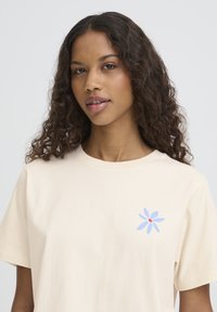 Kremno bela bombažna majica z okroglim izrezom, z modrim cvetličnim motivom z rdečim središčem na levi strani prsnega koša.