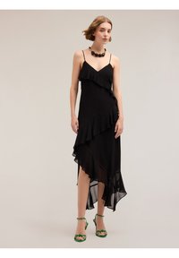 Rochie din chiffon negru cu un design stratificat de volane și margine asimetrică. Modelul poartă sandale verzi cu barete și un colier cu mărgele.