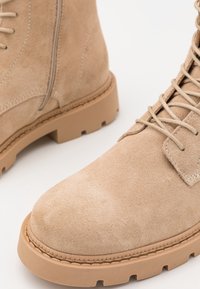 Stivaletti in suede color marrone chiaro con lacci, suole in gomma spesse e cerniere laterali su uno sfondo bianco.
