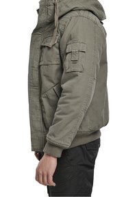 Zielona kurtka z kapturem, z przednimi kieszeniami, naszywaną kieszenią na rękawie, ściągaczami na mankietach i luźnym krojem.