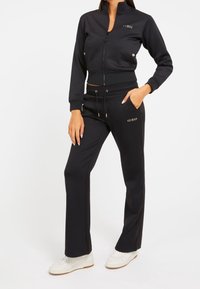 Kvinde iført en sort zip-up jakke og matchende sorte bukser med "GUESS" logo, parret med hvide sneakers, stående med en hånd i lommen.