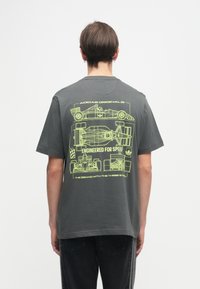 Γκρίζο βαμβακερό t-shirt με φωσφοριζέ κίτρινο γραφικό σχέδιο αγωνιστικών αυτοκινήτων και κείμενο "Σχεδιασμένο για Ταχύτητα" στην πλάτη. Κοντά μανίκια, στρογγυλή λαιμόκοψη.