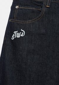 Jean en denim bleu foncé avec surpiqûres orange et petit logo blanc brodé près de la poche avant.