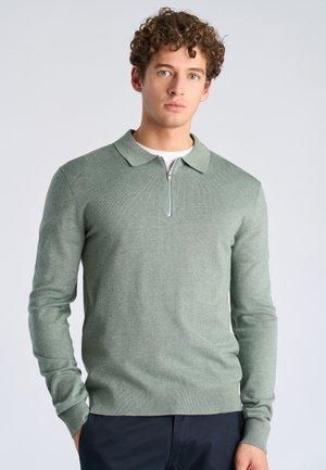 Młody mężczyzna o kręconych włosach, ubrany w zielony sweter z zamkiem i ciemne spodnie, stojący na tle jednolitego jasnego tła.