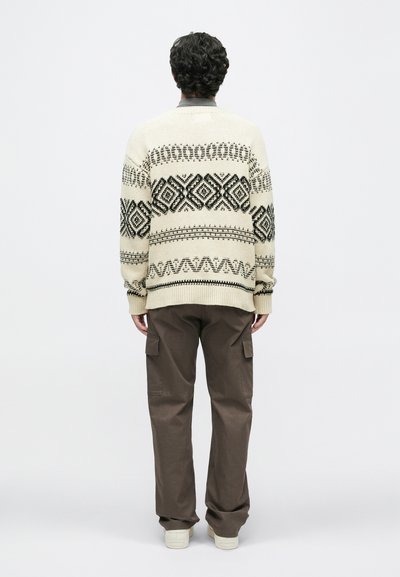 Beige kötött pulóver fekete geometrikus mintákkal, lazán szabott barna cargo nadrággal és világos színű lábbelikkel párosítva.
