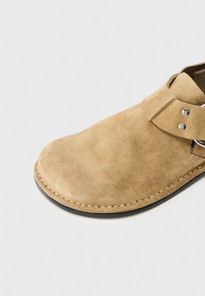 Παντόφλα από σουέντ σε ανοιχτό καφέ χρώμα με καμπυλωτή μύτη, επίπεδη σόλα, πλαϊνό λουράκι και μεταλλικές λεπτομέρειες. Λεπτομερής ραφή κατά μήκος της άκρης.