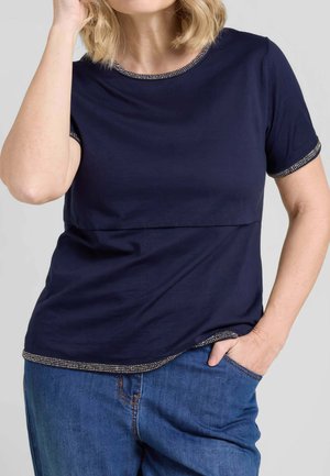 Donna con top blu navy a maniche corte con bordo di perline e jeans blu, che posa con una mano in tasca e l'altra vicino ai capelli.