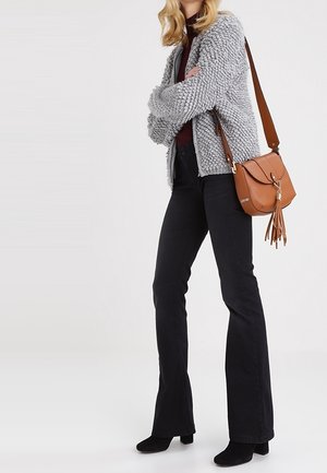 Žena v šedé texturované bundě, tmavých rozšířených kalhotách, černých botách a nosící hnědou crossbody tašku s detailním třásňovým zdobením.