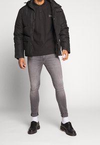 Chaqueta de plumas negra con capucha, combinada con una camiseta tipo polo oscura, jeans de corte slim gris claro y zapatos negros con calcetines blancos.