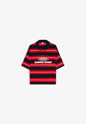 Rövid ujjú galléros póló piros és fekete vízszintes csíkokkal, fekete gallérral, és fehér "Surface Studio" logóval, amely kör alakú mintával díszített a mellkason.