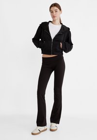 Crna skraćena hoodie majica sa prednjim patentnim zatvaračem, uparena s crnim širokim tajicama. Model nosi bijelu majicu i bež tenisice s smeškastim potplatom.