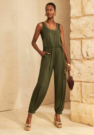 Vrouw die een olijfgroene mouwloze jumpsuit met kwastjesriem draagt, een kleine geweven tas vasthoudt en staat bij een gestructureerde stenen muur.