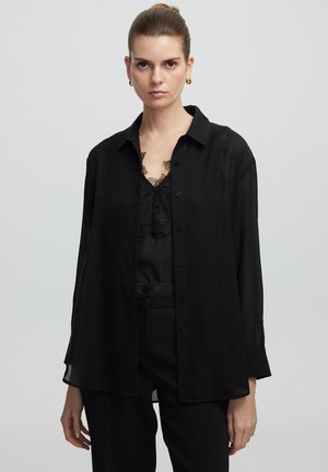 Chemise noire semi-transparente à boutons avec col, manches longues et coupe décontractée, superposée sur un top en dentelle noire. Texture lisse et tissu léger.