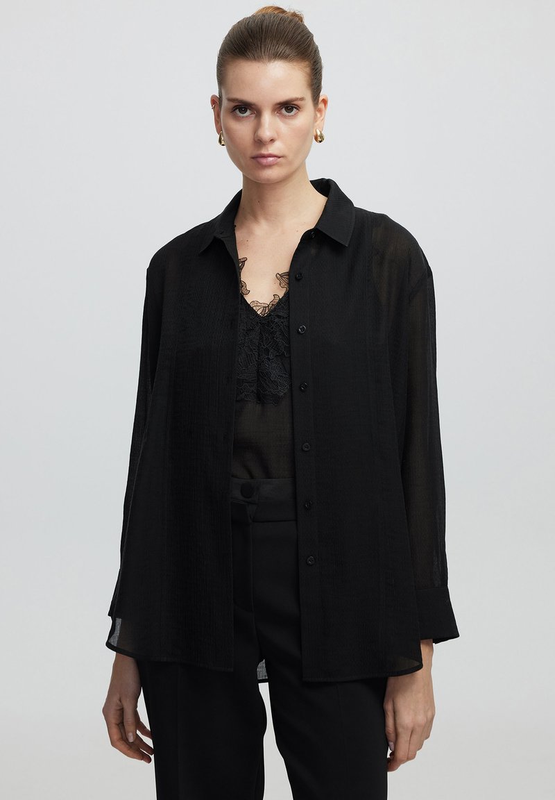 Camicia nera trasparente con bottoni, colletto, maniche lunghe e vestibilità rilassata, indossata sopra un top in pizzo nero. Tessuto liscio e leggero.