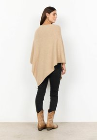 Poncho bej dintr-un material tricotat moale, având un design fluid cu tiv asimetric. Asortat cu blugi negri slim și bocanci maro de tip cowboy.