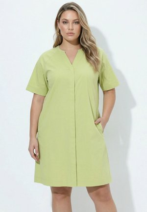 Frau mit langen blonden Haaren, die ein kurzärmeliges, hellgrünes, knielanges Kleid mit V-Ausschnitt und Seitentaschen trägt.