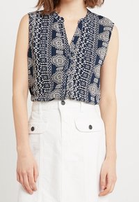 Blouse sans manches bleu marine avec une patte de boutonnage sur le devant, présentant un motif géométrique et cachemire crème. Portée avec une jupe blanche taille haute.