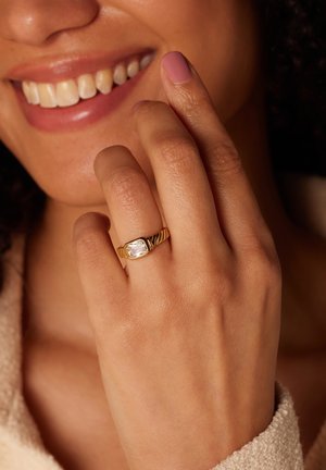 Gouden ring met een grote heldere steen, voorzien van een gedraaide bandontwerp en een gepolijste afwerking. Hand rust tegen een zachte, gestructureerde ondergrond.