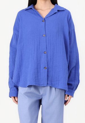 Femme portant une chemise bleue ample à manches longues avec col et un pantalon bleu clair, sur un fond uni.