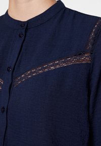 Blusa blu navy con colletto alla cinese, chiusura a bottoni e dettagli in pizzo intricati sul petto. Tessuto morbido con una finitura testurizzata.