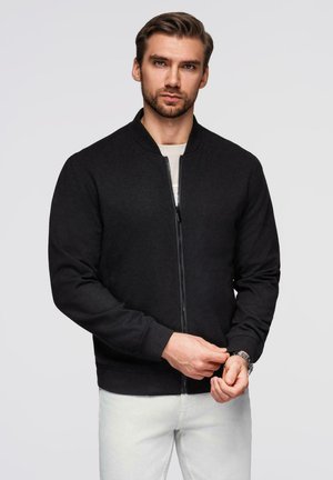 Veste bomber noire zippée avec des poignets et un col côtelés, fabriquée dans un tissu doux. Le mannequin porte un pantalon de couleur claire, mettant en avant un style décontracté.